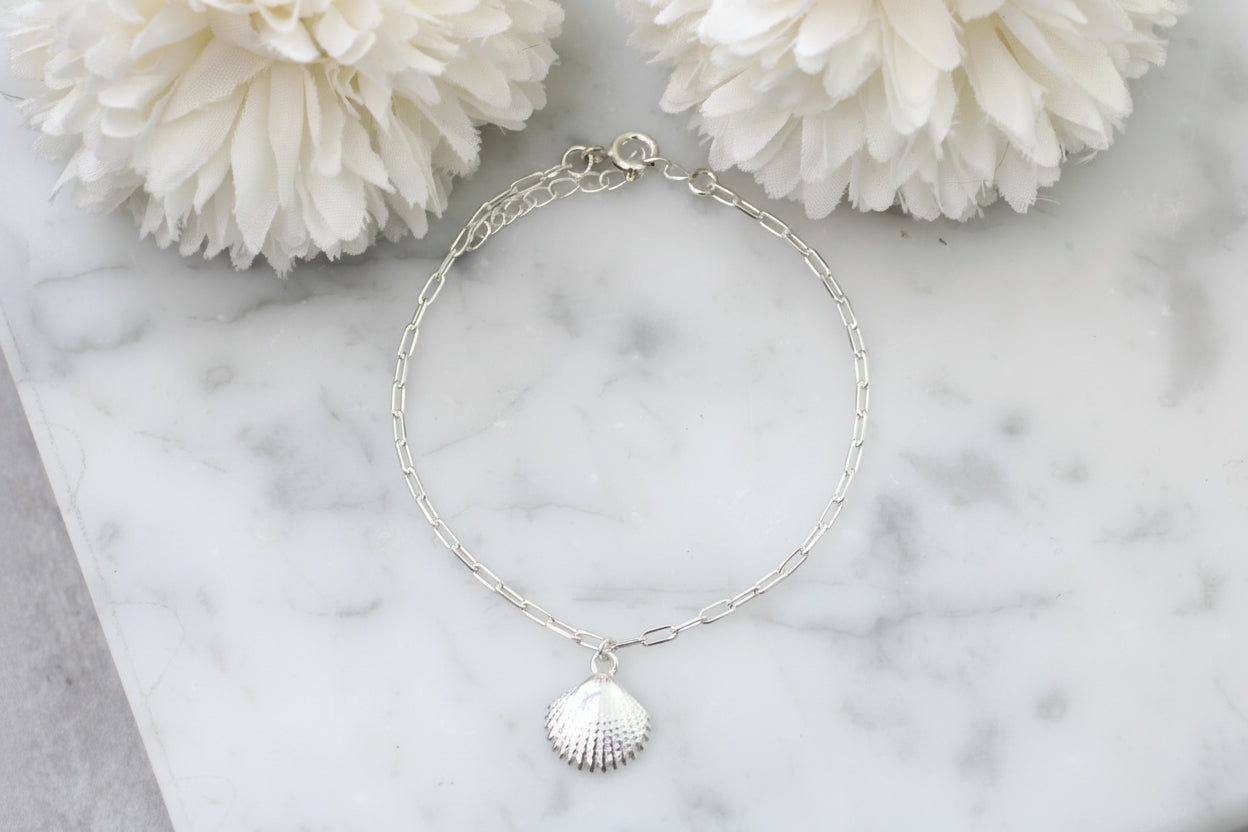 Silver anklet with a shell charm on a marble surface with white flowers.