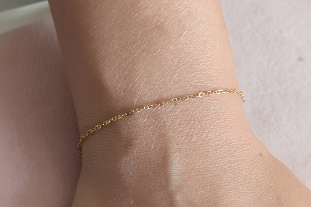 A delicate gold chain bracelet worn on a person's wrist, shown in a close-up view against a soft pink background.