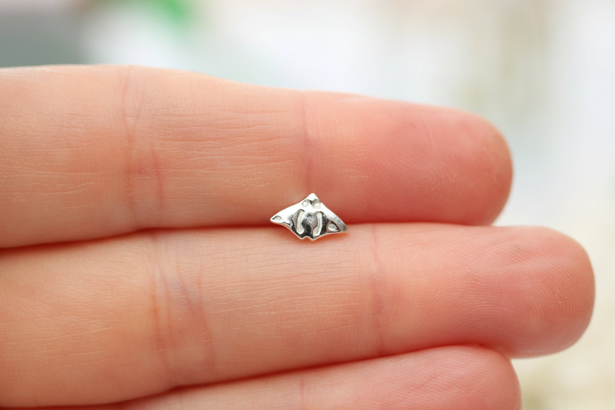 A tiny silver-toned metal charm or bead with a small raised circular detail, resting on a person's fingers against a soft blurred light background.