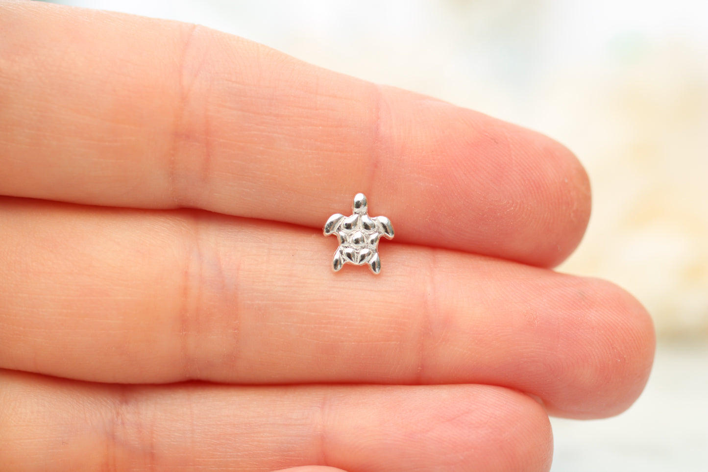 A tiny silver sea turtle charm resting on the fingertips of a hand, shown in close-up to demonstrate its small scale.