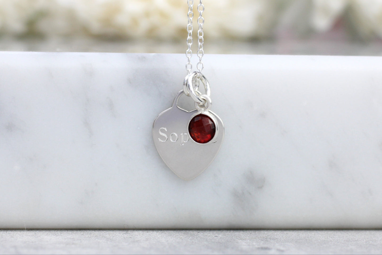 Silver necklace with a heart-shaped pendant featuring a red gemstone and engraved text, on a marble surface.