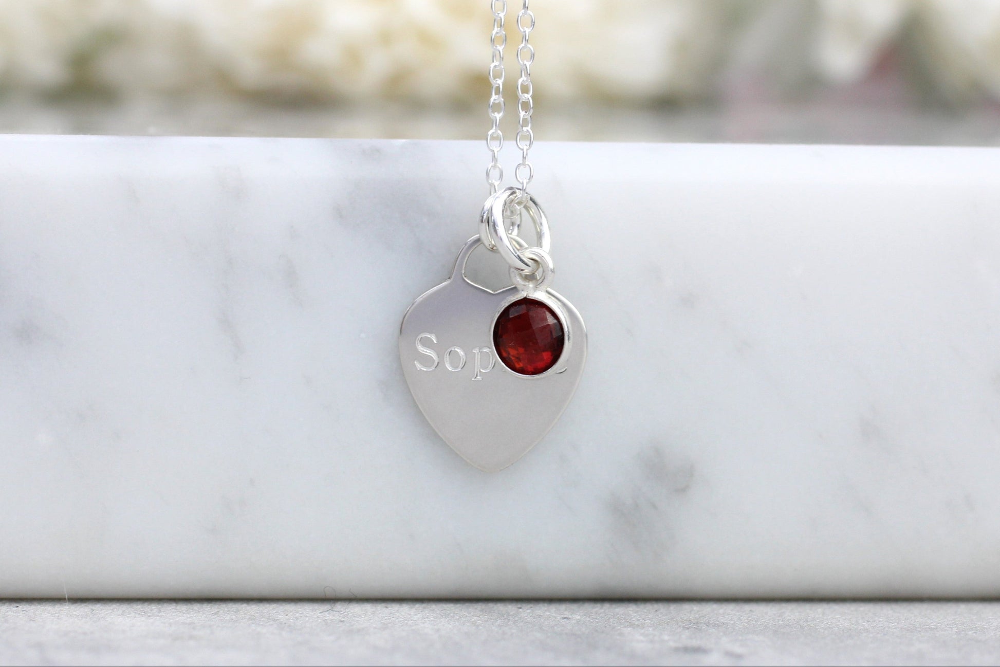 Silver necklace with a heart-shaped pendant featuring a red gemstone and engraved text, on a marble surface.
