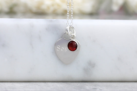 Silver necklace with a heart-shaped pendant featuring a red gemstone and engraved text, on a marble surface.