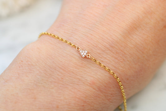 Gold bracelet with a heart-shaped charm on a wrist against a neutral background