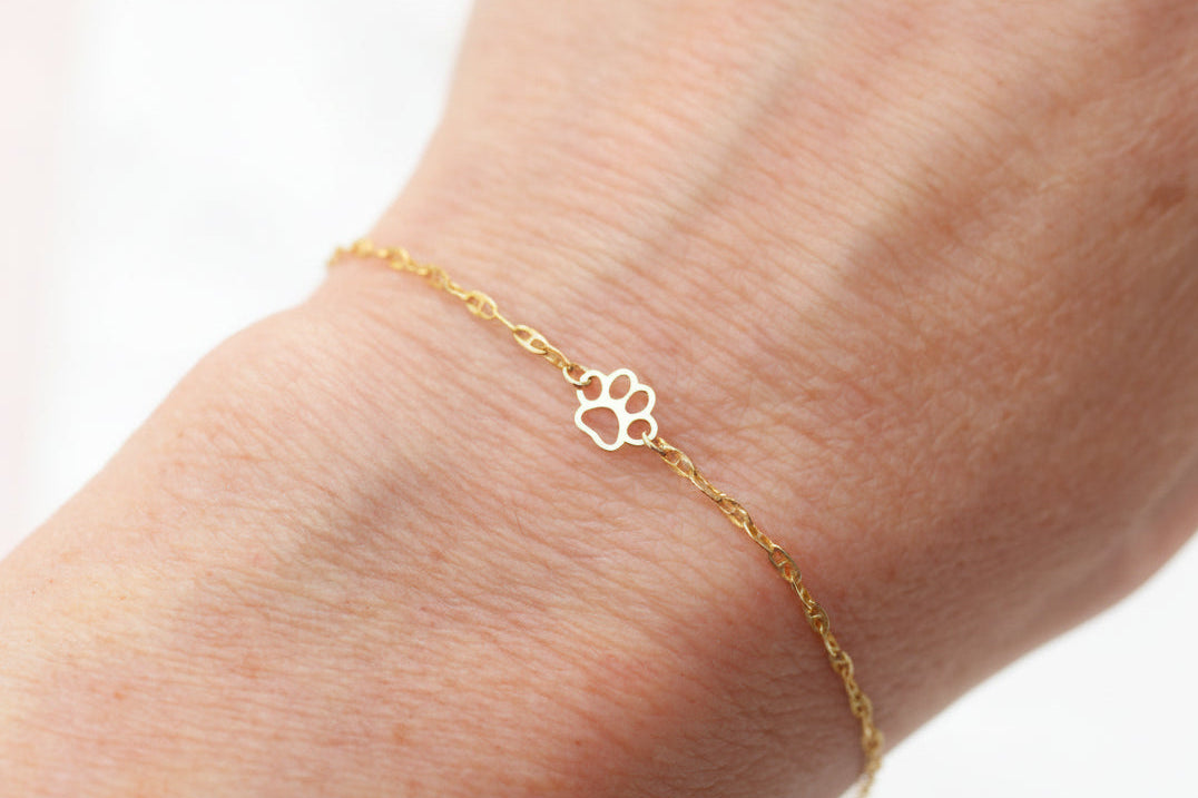 A delicate gold chain bracelet with a small open paw print charm, shown worn on a person's wrist against a soft white background.