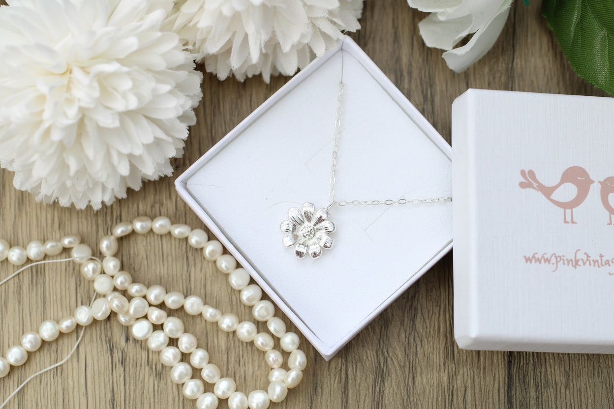 Silver necklace with a flower pendant on a white jewelry box, surrounded by pearls and white flowers on a wooden surface.
