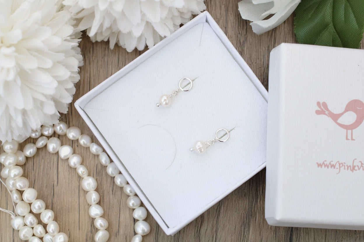 Pearl earrings in a white box with a pink bird logo on a wooden surface.