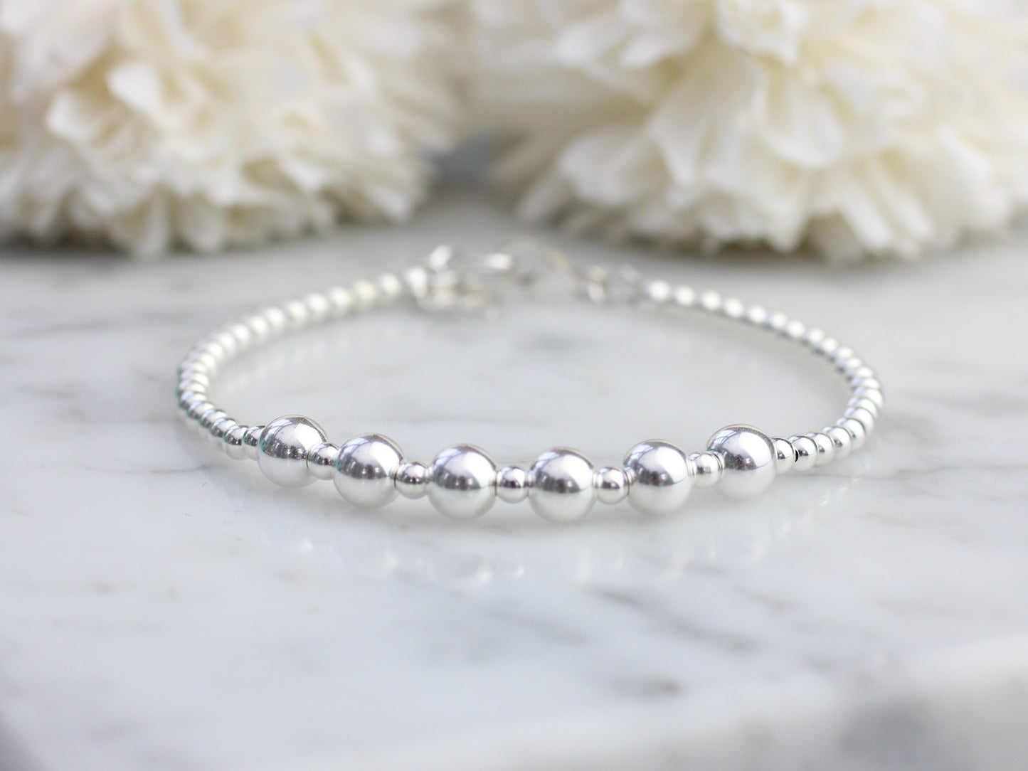 Silver beaded bracelet on a marble surface with white flowers in the background
