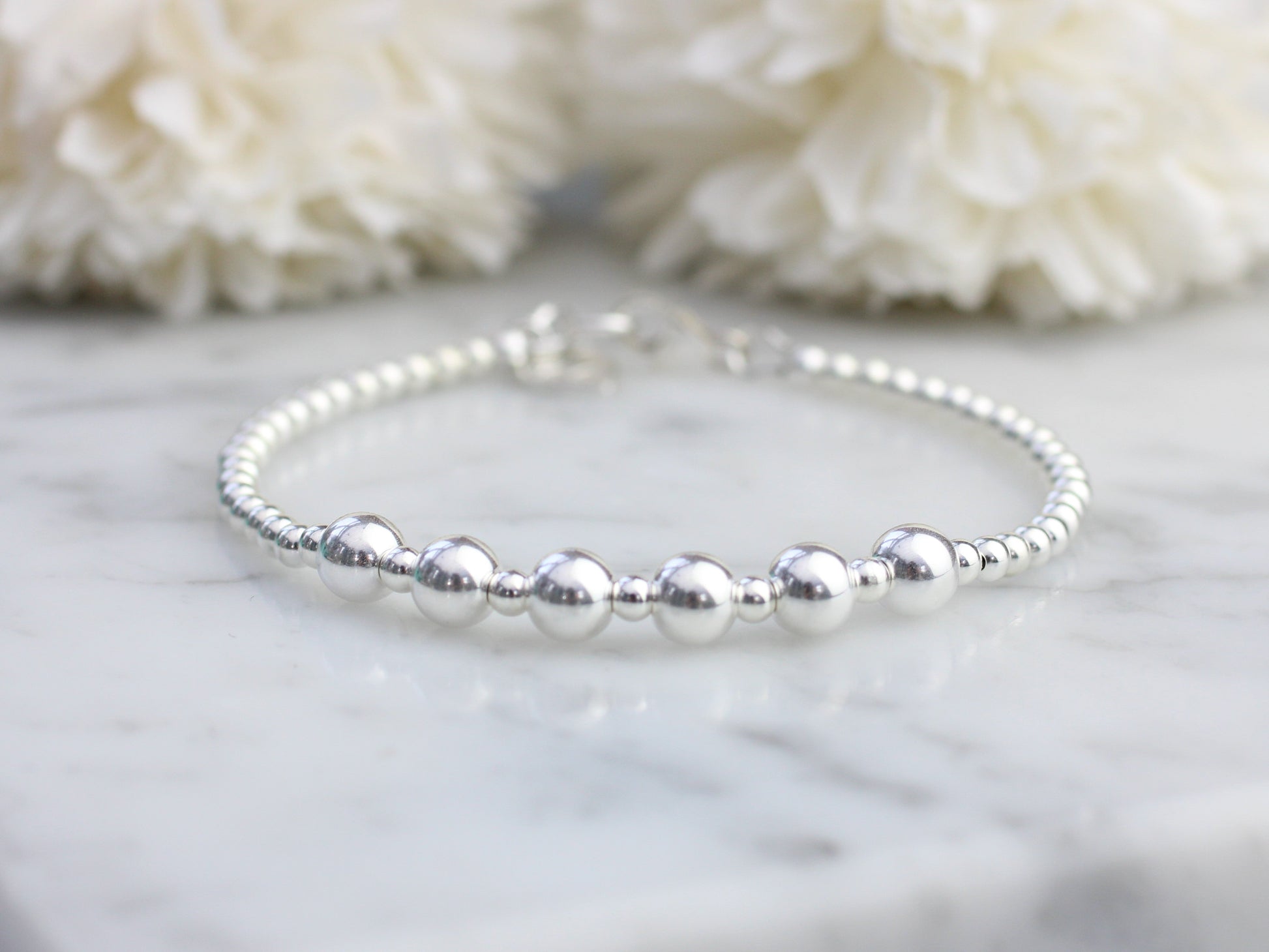 Silver beaded bracelet on a marble surface with white flowers in the background