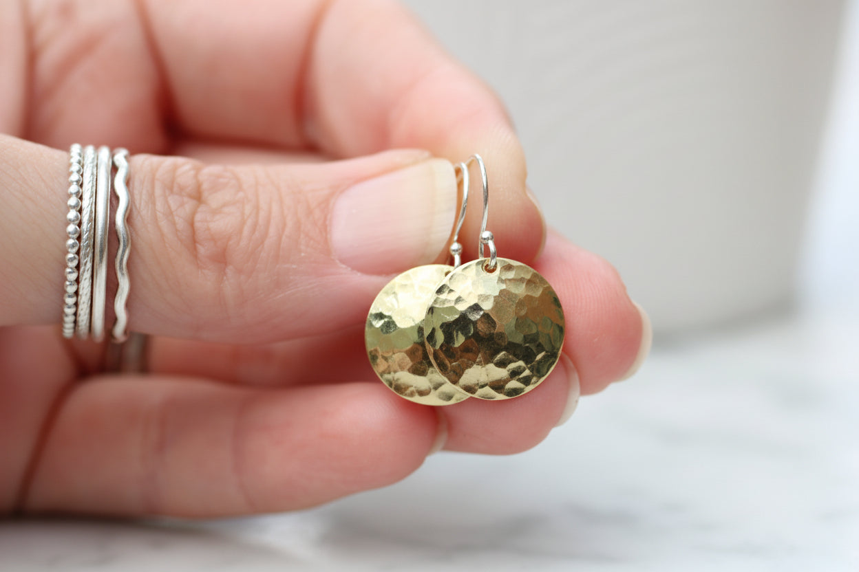 A close-up of a hand holding a pair of hammered gold oval dangle earrings with silver-toned hooks. The hand has several silver rings on the fingers, and the background is a soft, out-of-focus light gray and white setting.