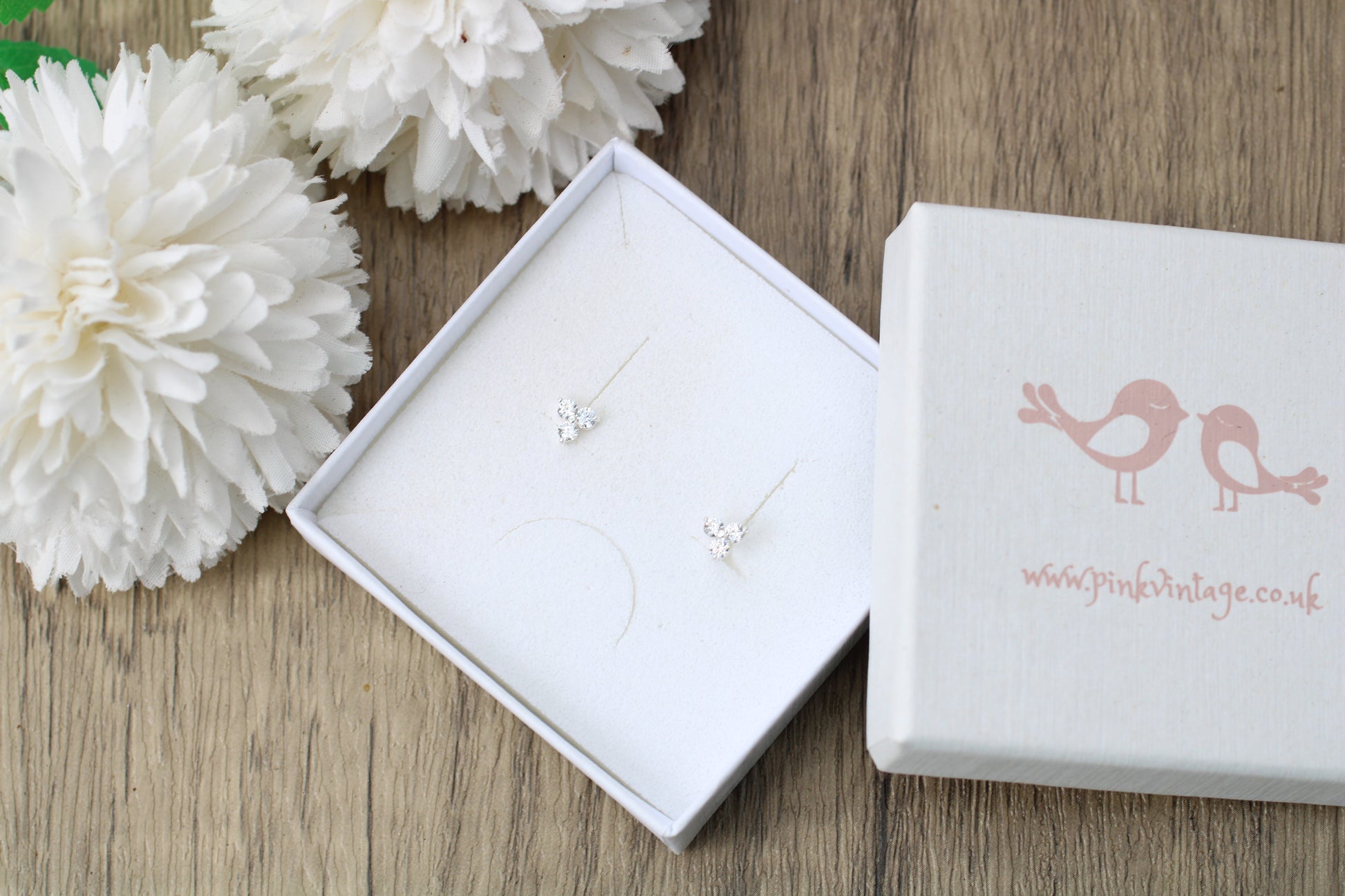 A pair of delicate silver stud earrings displayed in an open white jewelry box next to a closed white gift box printed with two pink birds and the website www.jpinkvintage.
