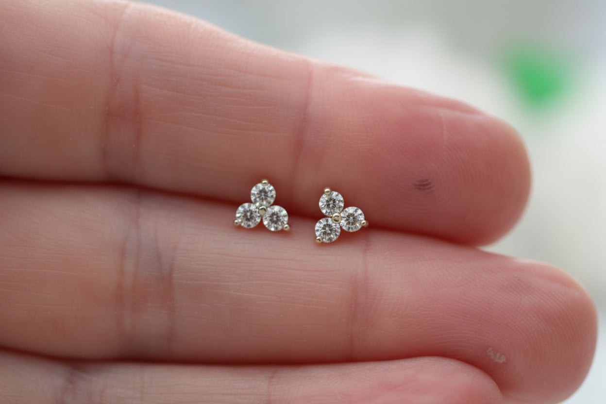 A close-up of a person's hand holding a pair of small silver stud earrings with clear gemstones, set against a softly blurred light background.