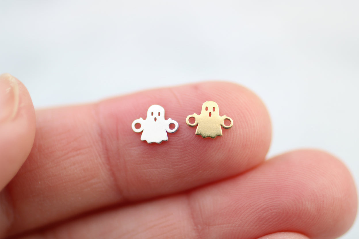 Two small ghost-shaped charms, one white and one gold, held between fingers on a white background.