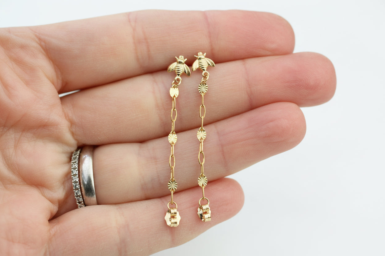 Gold bee-shaped earrings held between fingers with a blurred background
