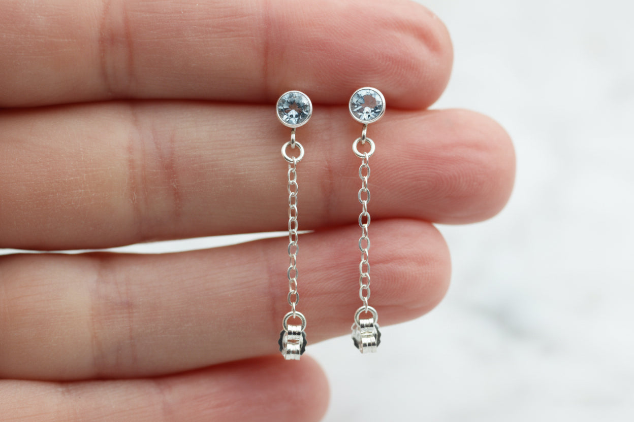 Silver chain earrings with gemstones held between fingers against a neutral background