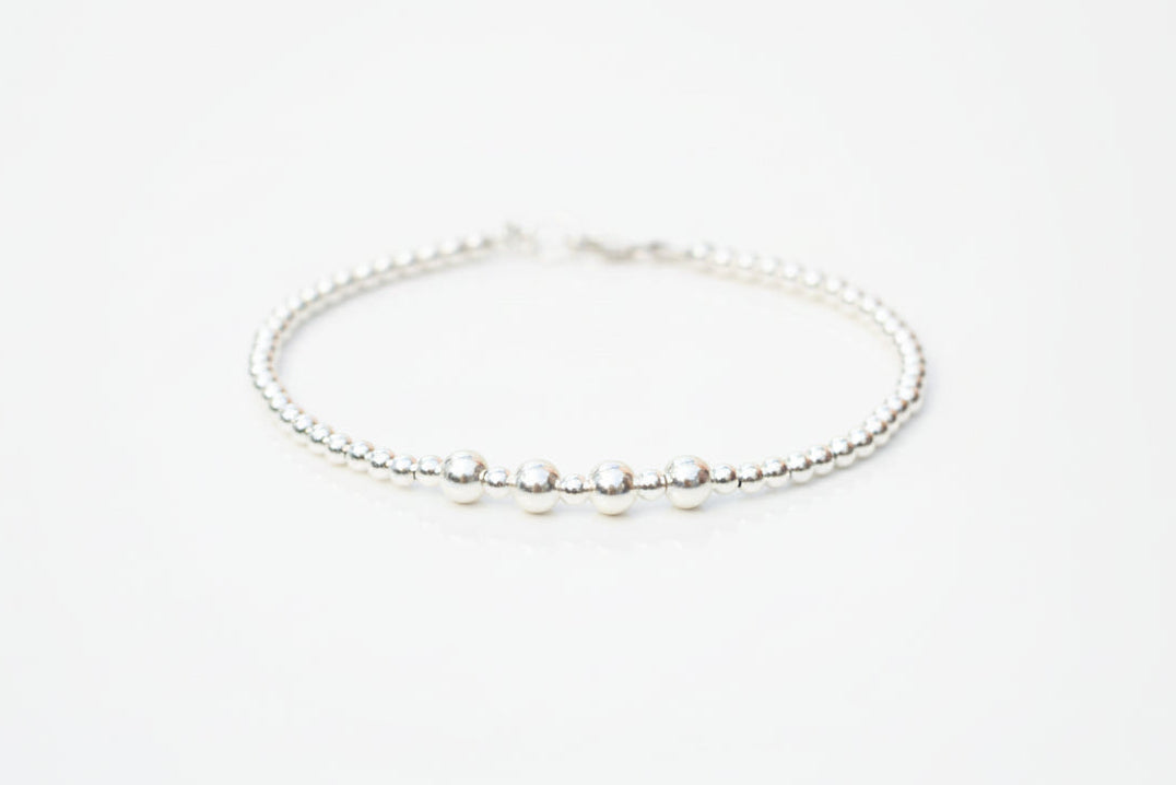 A delicate silver beaded bracelet with four larger polished silver beads, displayed on a white surface with soft-focus white flowers in the background.