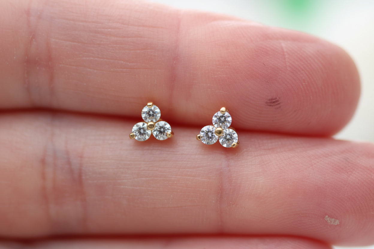 A pair of small gold stud earrings with three clear round-cut stones arranged in a triangle, shown resting on a person's fingers.