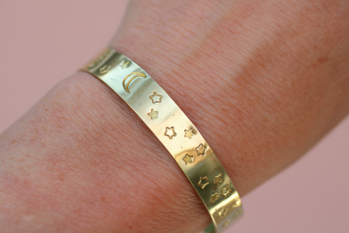 A close-up of a gold-toned metal cuff bracelet with engraved crescent moon and star patterns, worn on a person's wrist.