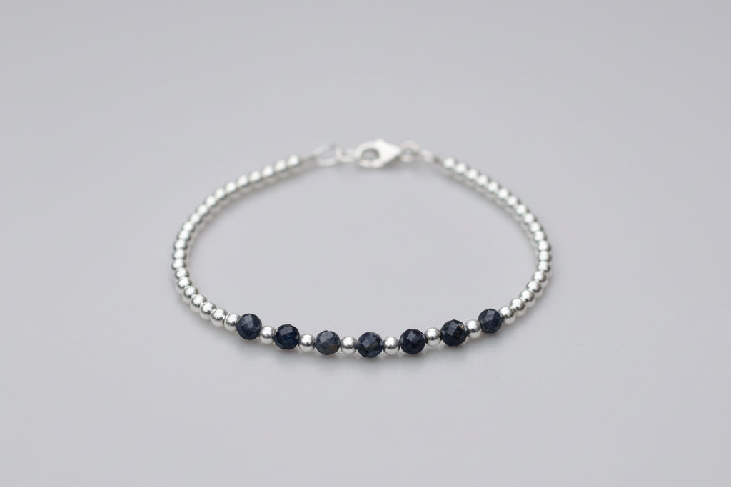 A delicate silver beaded bracelet with five dark navy blue beads, displayed on a white marble surface with blurred white flowers in the background.