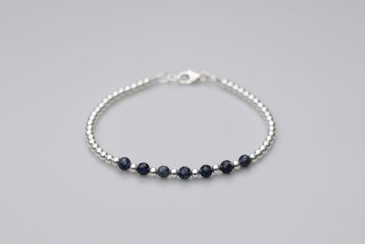 A delicate silver beaded bracelet with five dark navy blue beads, displayed on a white marble surface with blurred white flowers in the background.