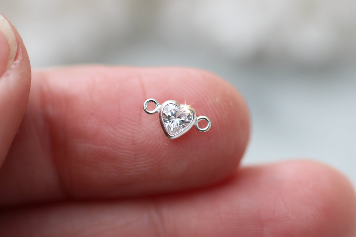 A tiny silver heart-shaped charm with a clear faceted gemstone in the center, held between two human fingers to show its small scale.