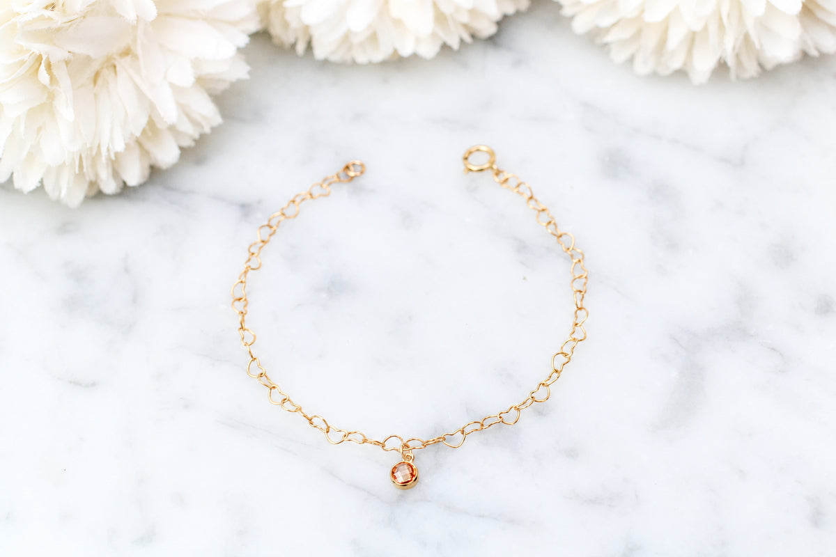 A delicate gold chain bracelet with a small rose gold oval charm, displayed on a white marble surface with white flowers in the background.