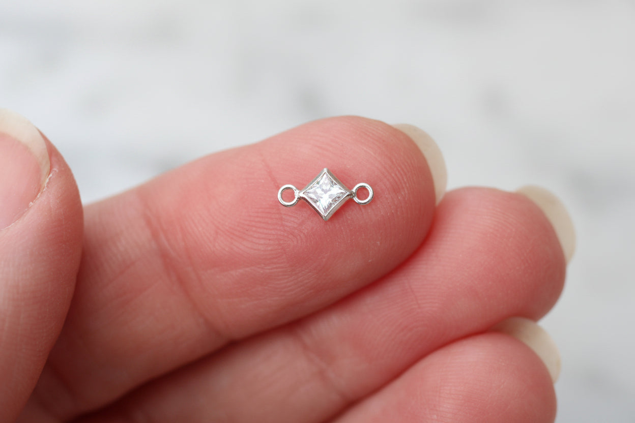 A tiny silver diamond-shaped jewelry charm with a clear faceted stone in the center, held between two fingers against a soft, blurred light gray background.