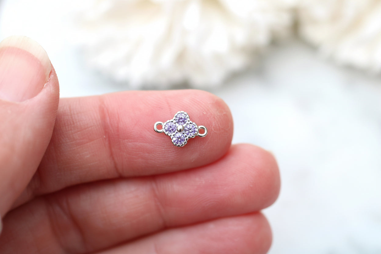 A small silver-toned four-petal charm with a purple center, held on a person's fingers against a soft white background.