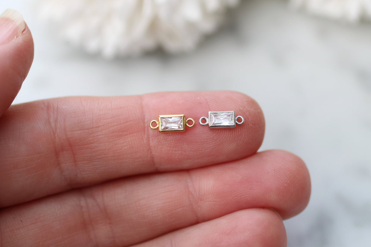 Two small rectangular clear gemstone jewelry findings with gold and silver settings, held on a person's fingers against a soft white marble background.