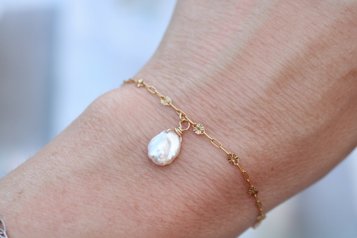 A delicate gold chain bracelet with small floral motifs and a teardrop-shaped white baroque pearl charm, shown worn on a person's wrist.