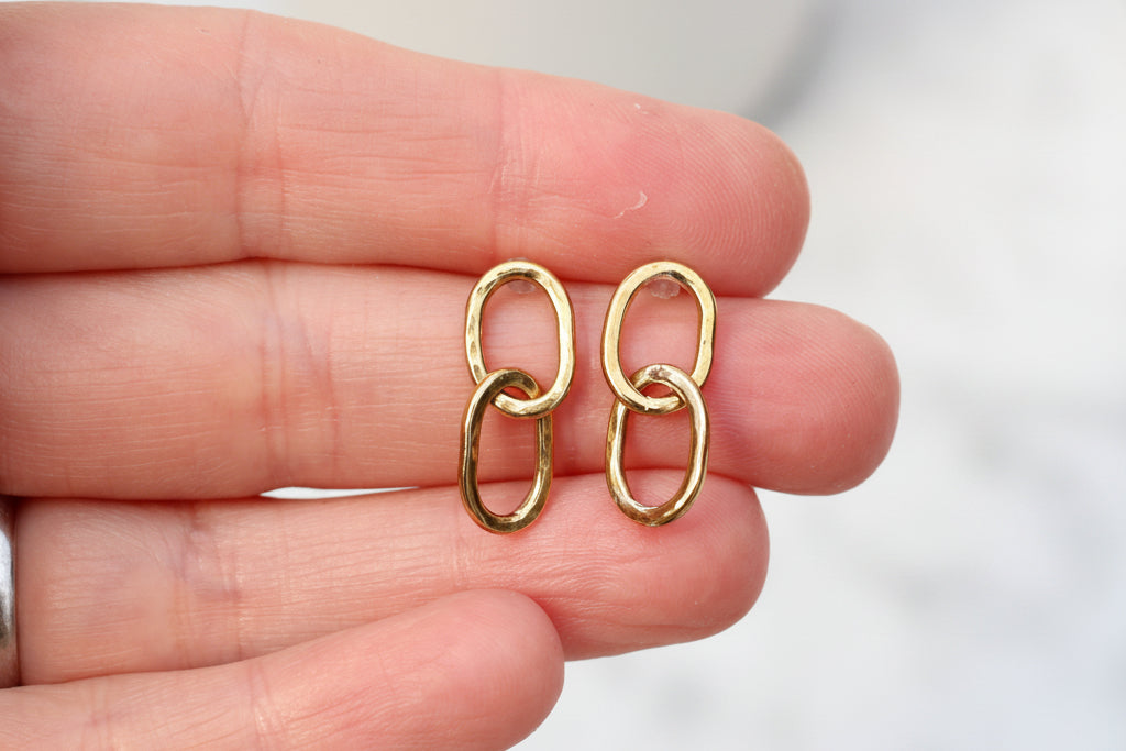 A pair of small gold link earrings held between a person's fingers against a soft, blurred light background.