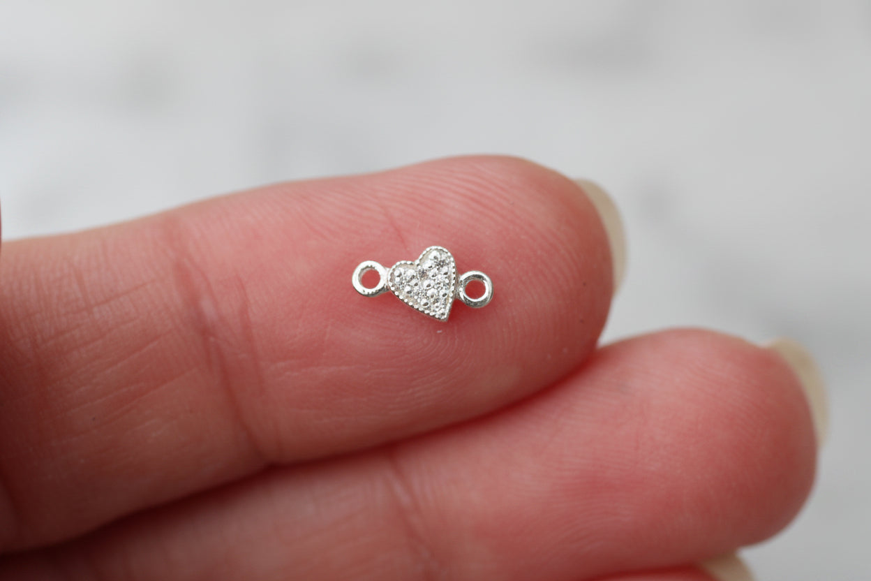 A tiny silver heart charm with clear rhinestones, held between two fingers to show its small scale.