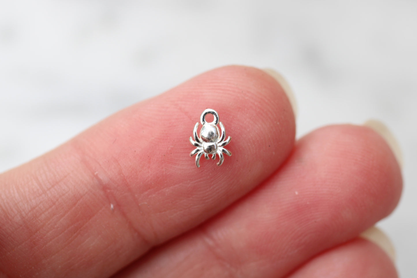 A tiny silver spider charm with a small loop at the top, held between two fingers to show its small scale.