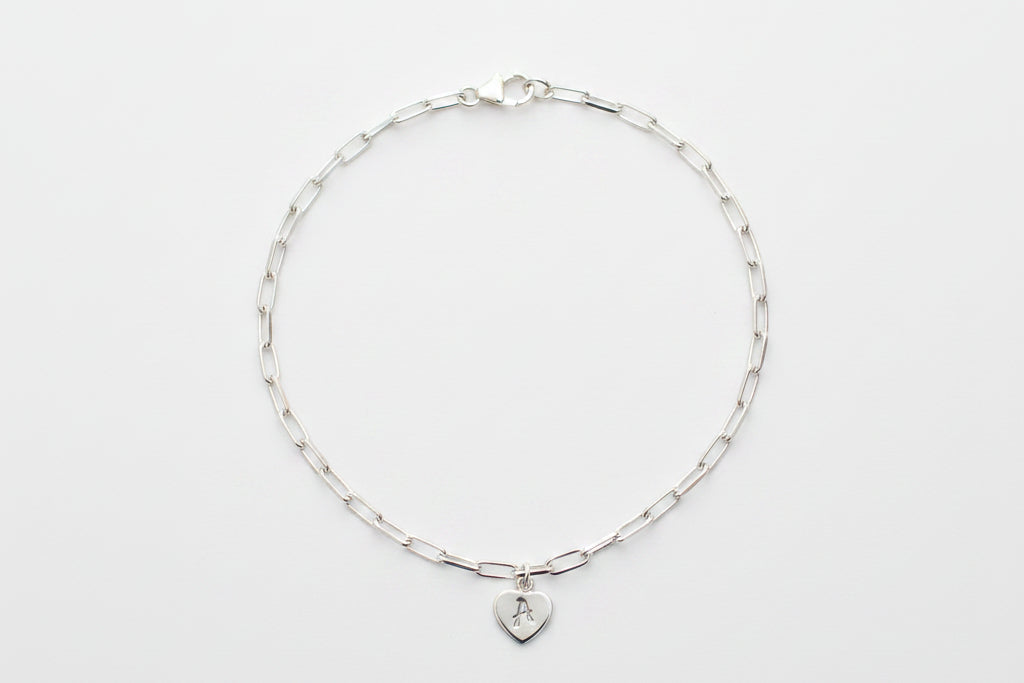 A delicate silver paperclip chain bracelet with a small heart-shaped pendant engraved with the letter 'A', displayed on a white and gray marble surface.