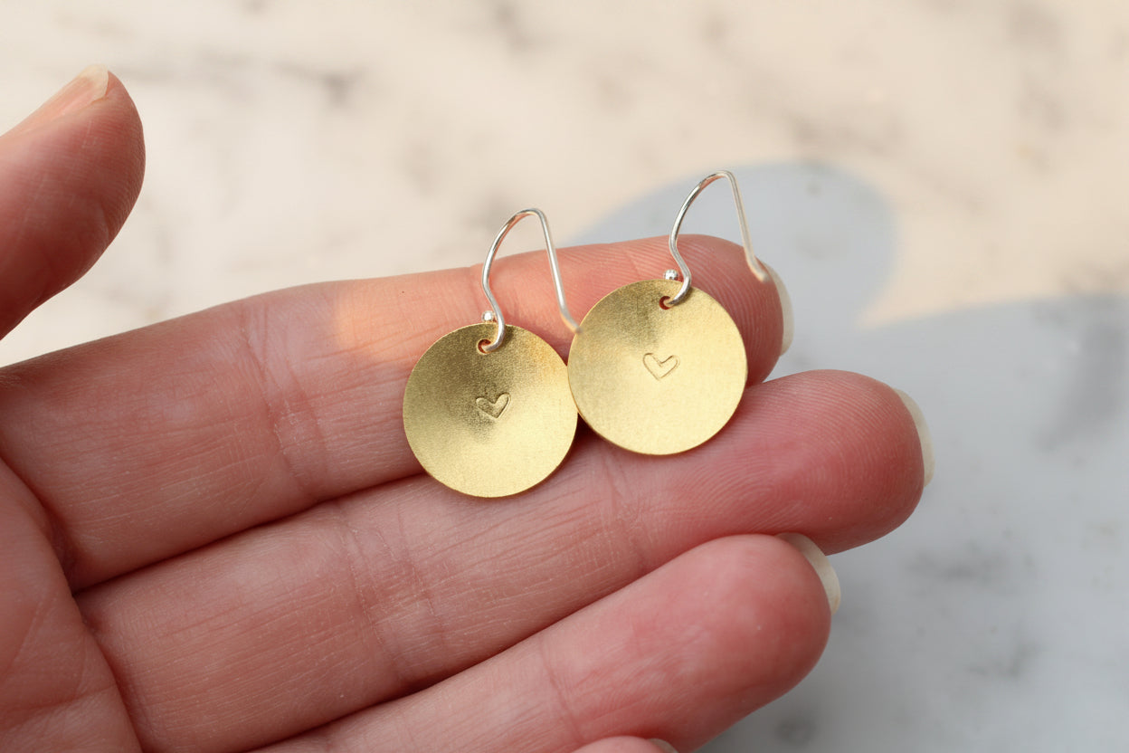A pair of gold disc drop earrings with silver fishhook wires, held on a person's fingers against a light marble background.