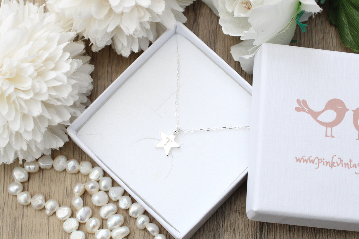 A delicate silver starfish necklace displayed in an open white gift box, surrounded by white pearls and white flowers on a wooden surface, with a branded white box lid next to it.