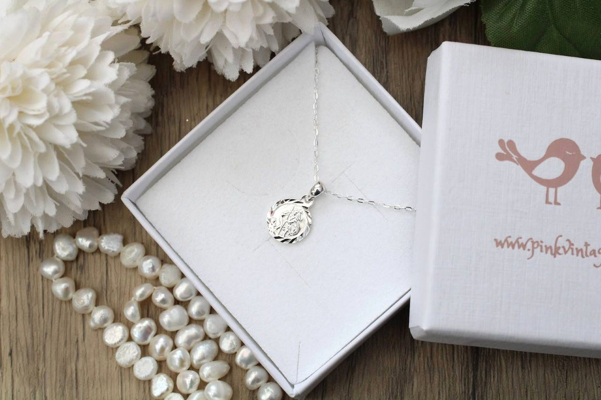 A silver pendant necklace with a circular engraved design, displayed inside an open white gift box alongside a branded white box lid and a strand of white pearls on a wooden surface.