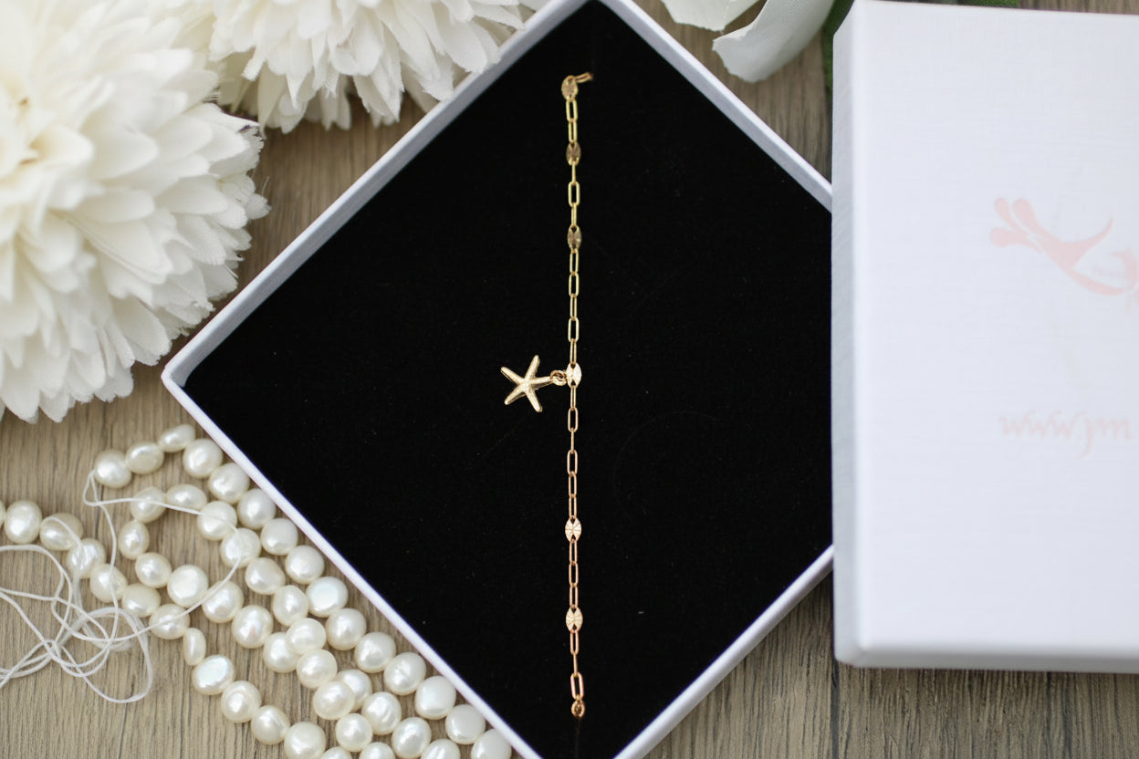 Gold bracelet with star charm in an open jewelry box on a wooden surface with flowers and pearls.