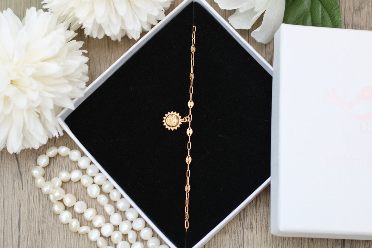 Gold anklet with sun design in a jewelry box on a wooden surface with flowers and pearls.