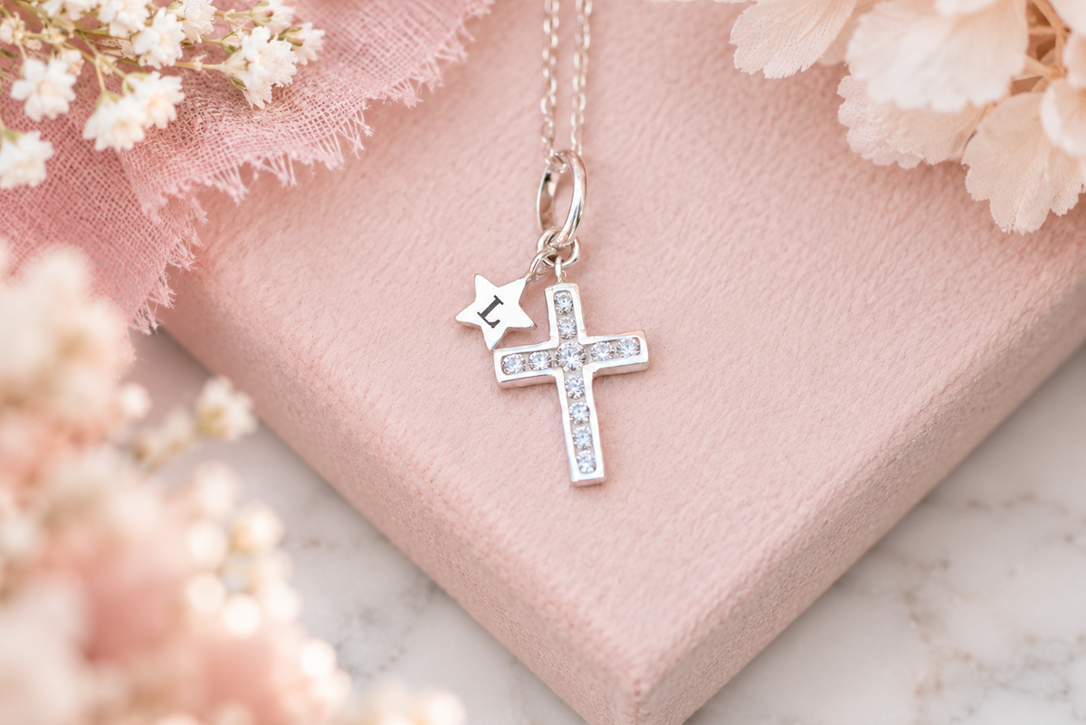 A silver cross necklace with a small star charm featuring the letter 'S', displayed on a soft pink gift box with delicate white flowers.