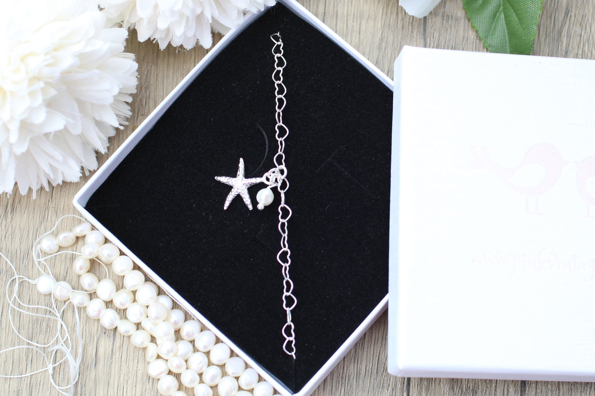 Silver anklet with starfish charm in a black jewelry box on a wooden surface with white flowers.