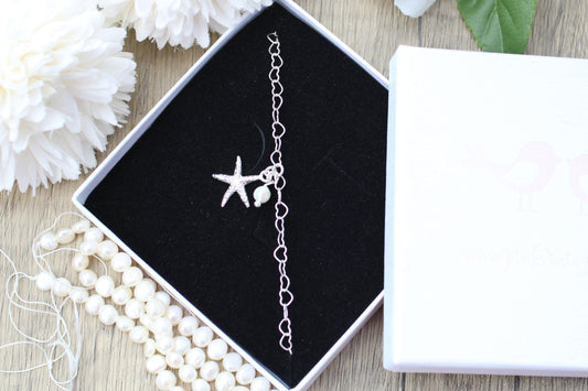 Silver anklet with starfish charm in a black jewelry box on a wooden surface with white flowers.