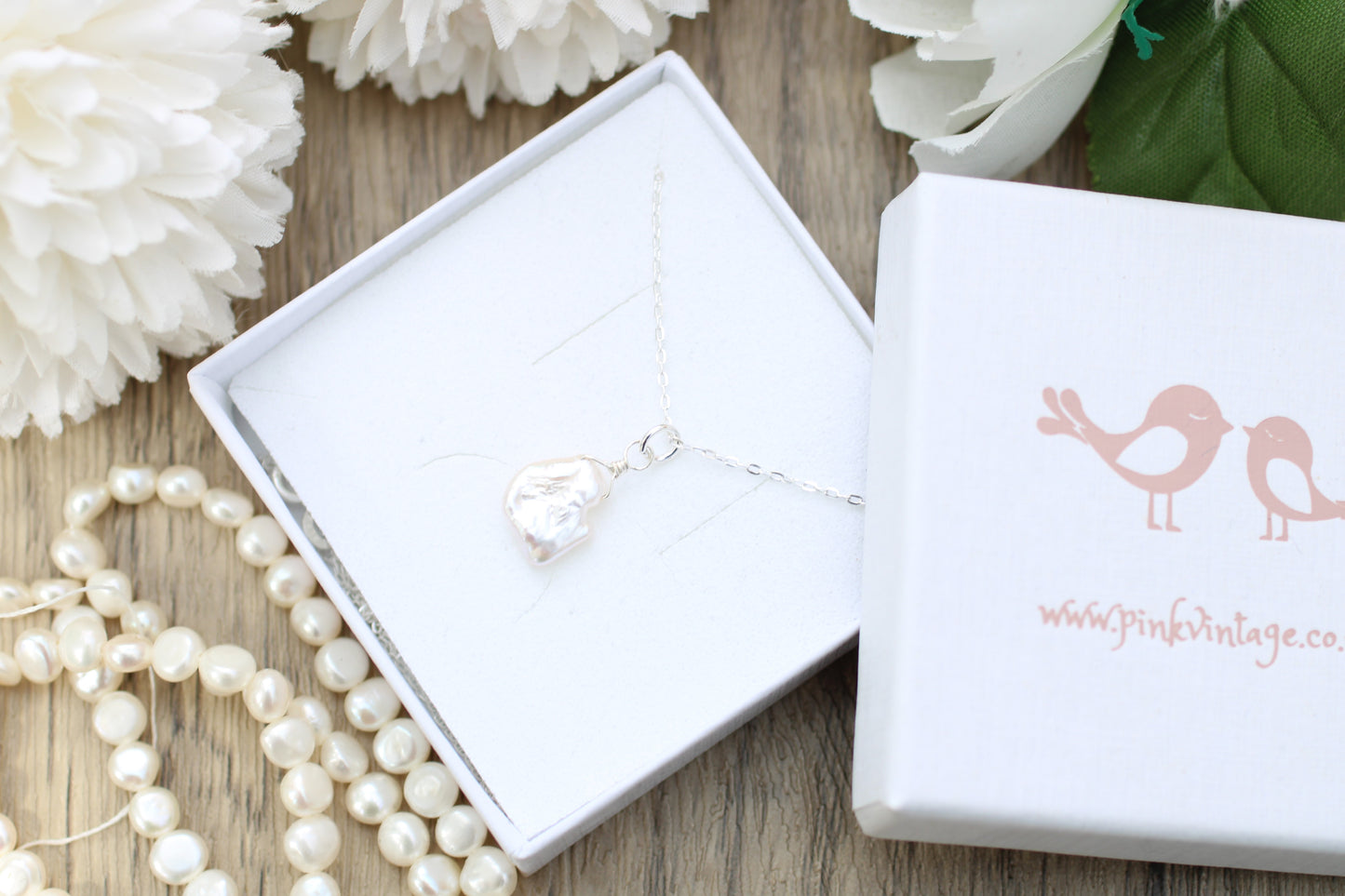 Silver necklace in a white box with 'Pink Vintage' branding, surrounded by pearls and flowers on a wooden surface.