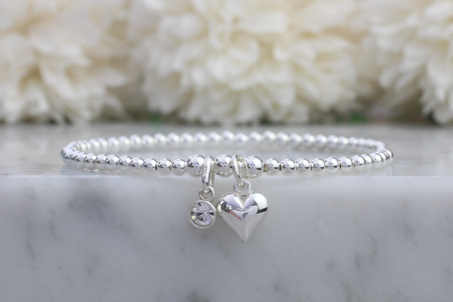 A sterling silver bracelet with a heart-shaped charm and a clear crystal or quartz gemstone, displayed on a marble surface.