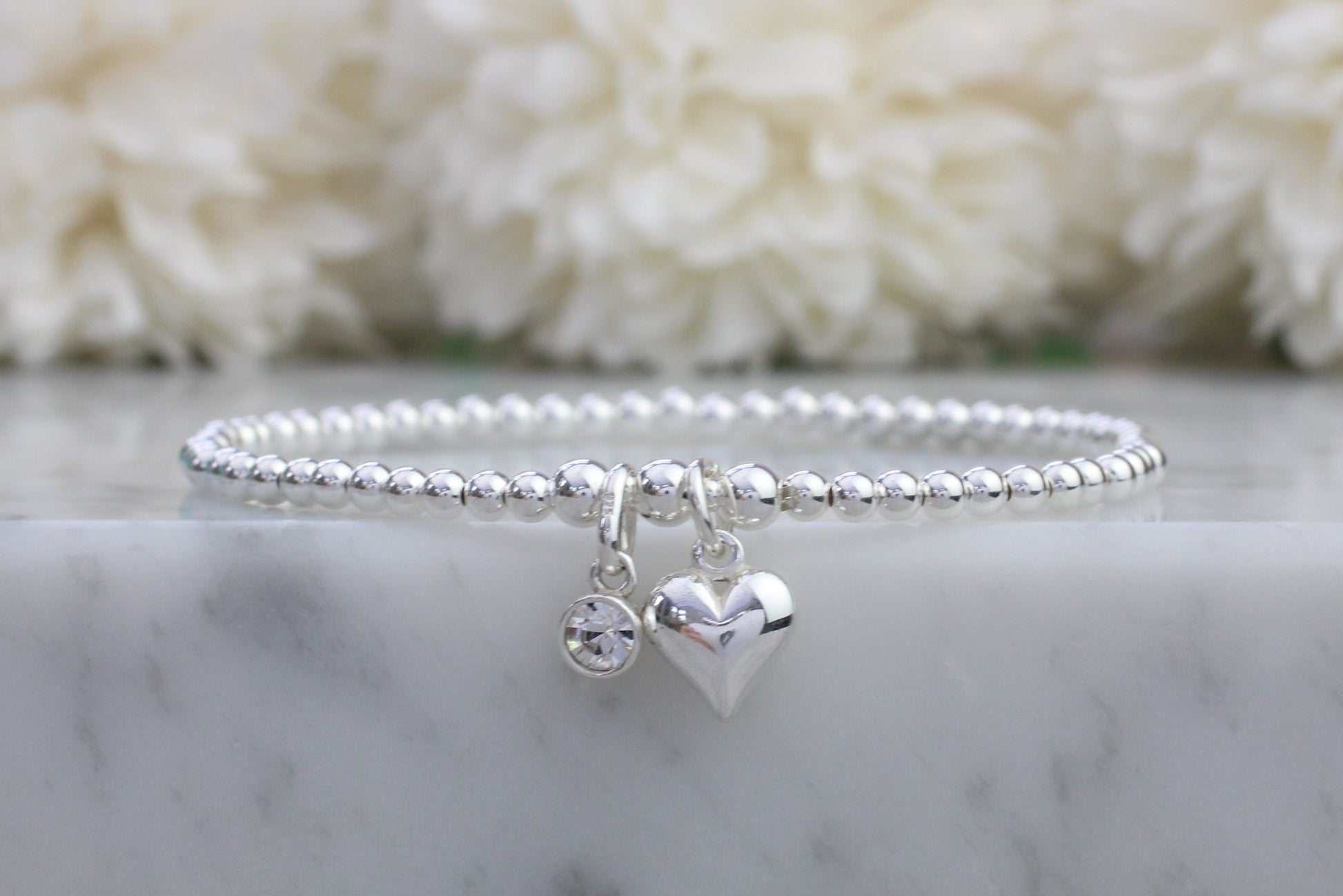 A sterling silver bracelet with a heart-shaped charm and a clear crystal or quartz gemstone, displayed on a marble surface.