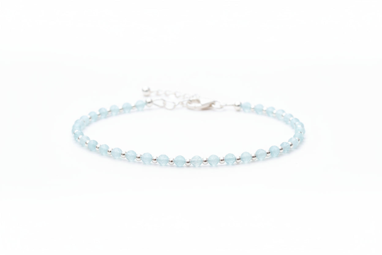 A silver bracelet with aquamarine gemstones laid out on a textured surface, with a jewellery box and white flowers in the background.