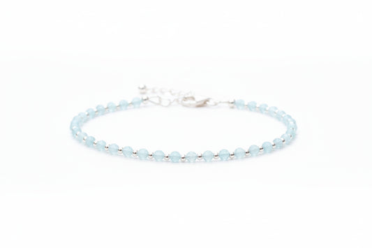 A silver bracelet with aquamarine gemstones laid out on a textured surface, with a jewellery box and white flowers in the background.