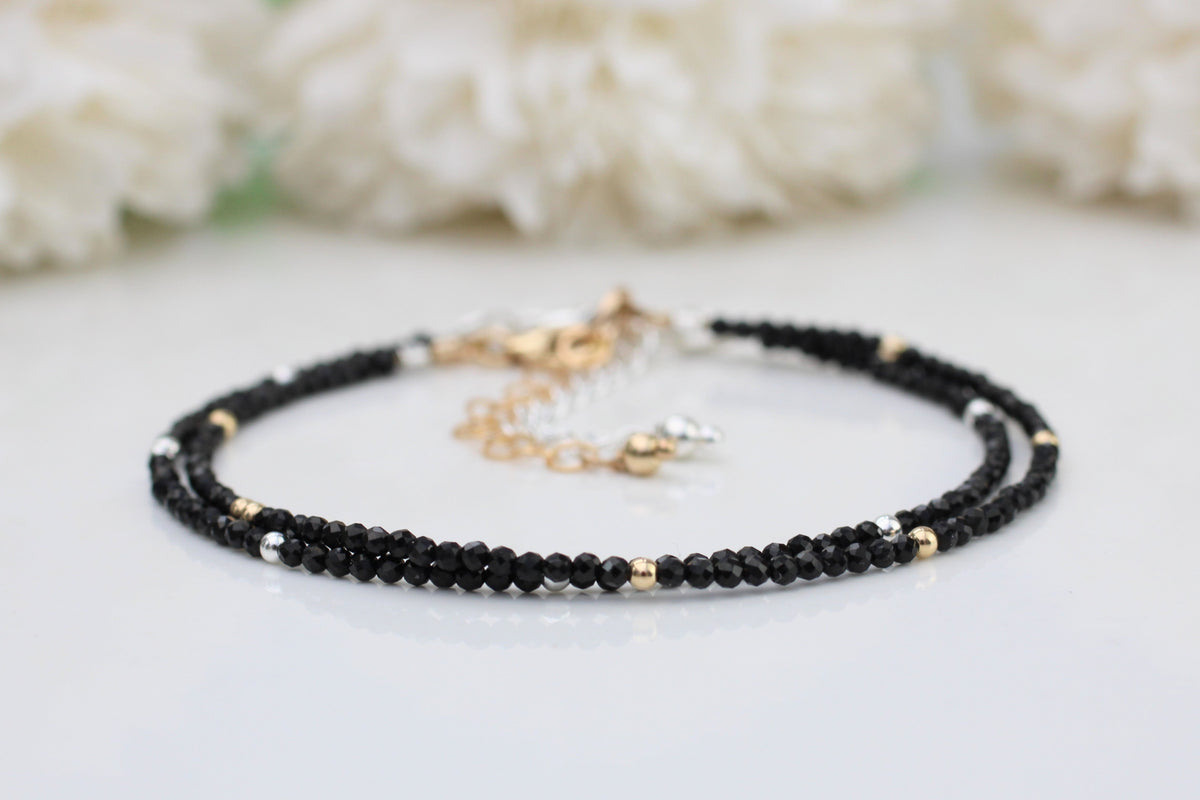 A black tourmaline bracelet with gold-filled metal accents, featuring 2mm gemstones and small beads, displayed on a white surface with white flowers in the background.