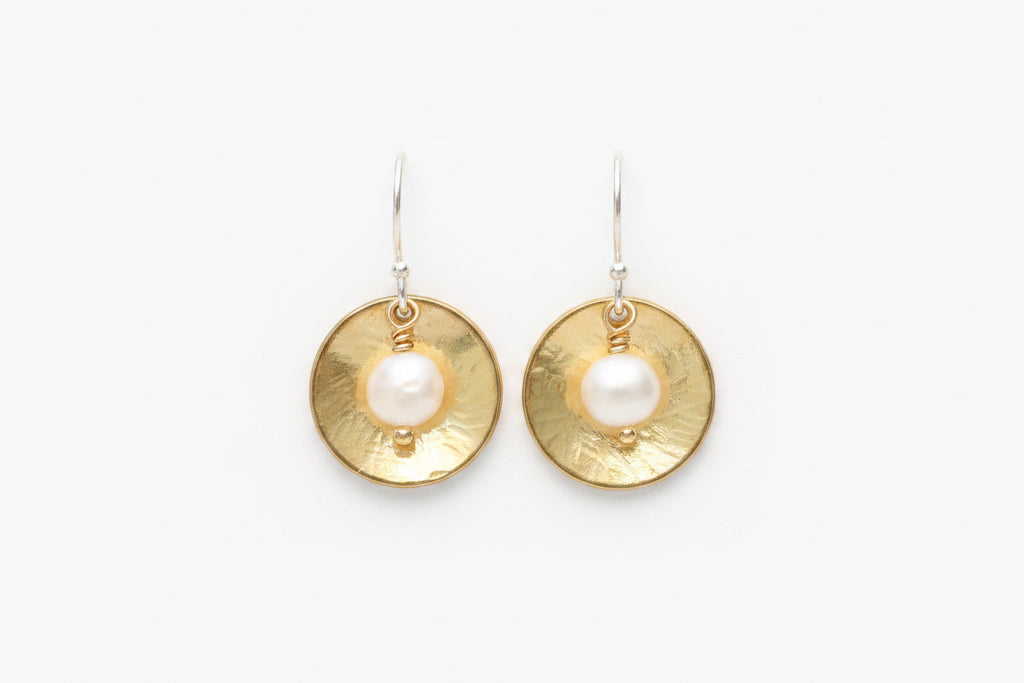 A pair of gold and silver drop earrings featuring a textured circular gold disc with a white pearl at the center, suspended from silver hooks on a plain white background.