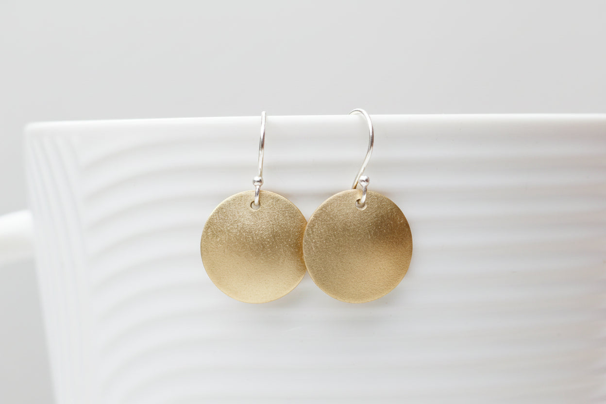 A pair of round gold-tone dangle earrings with silver fishhook wires, displayed on the rim of a white textured ceramic cup with a blurred background.